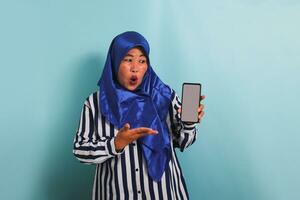 ein aufgeregt mittleren Alters asiatisch Frau im ein Blau Hijab und ein gestreift Hemd ist zeigen ein Telefon mit ein leer Weiß Bildschirm. sie ist isoliert auf ein Blau Hintergrund foto