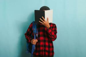 ein jung asiatisch Mann ist Abdeckung seine Gesicht mit ein Buch während Stehen gegen ein Blau Hintergrund foto
