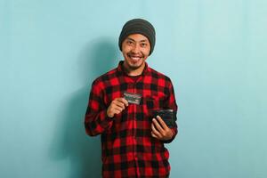 aufgeregt jung asiatisch Mann mit Mütze Hut und rot Plaid Flanell Hemd halten ein Brieftasche und Anerkennung Karte, isoliert auf ein Blau Hintergrund foto