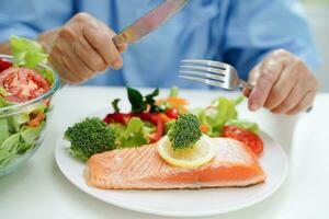 asiatisch Alten Frau geduldig Essen Lachs Anteil und Gemüse Salat zum gesund Essen im Krankenhaus. foto