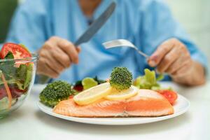 asiatisch Alten Frau geduldig Essen Lachs Steak Frühstück mit Gemüse gesund Essen während Sitzung und hungrig auf Bett im Krankenhaus. foto