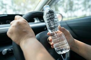 asiatische Fahrerin, die eine Flasche zum Trinken von Wasser hält, während sie ein Auto fährt. Wärmflasche aus Kunststoff verursacht Feuer. foto