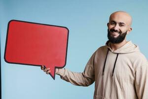 lächelnd arabisch Mann halten Rede Blase zum Produkt Werbung spotten oben Porträt. heiter jung Person posieren mit sprechen Ballon mit Kopieren Raum spotten oben und suchen beim Kamera foto