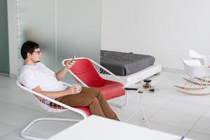 Mann sitzt auf einem Stuhl und schaut fern und hält Teetasse und Telefon in der Hand foto
