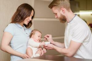 Vater und Mutter Futter ihr Baby Obst Püree im das Küche von ein Löffel foto