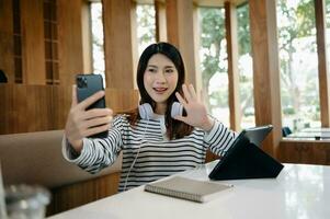 attraktiv jung asiatisch Schüler studieren beim das Hochschule Bibliothek, Sitzung beim das Schreibtisch, mit ein Laptop Computer, Tablette und Kopfhörer haben ein Video Plaudern. foto