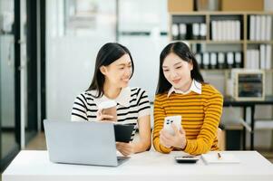 zwei attraktiv jung asiatisch weiblich Hochschule Studenten Arbeiten auf das Schule Projekt mit Laptop Computer und Tablette zusammen, genießen reden und Essen foto