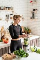 junge blonde lächelnde Frau, die zu Hause in der Küche grünen Smoothie macht foto