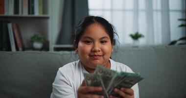 Porträt von jung asiatisch Frau genießen Zählen Kasse Dollar Banknoten auf Sofa im das Leben Zimmer beim heim. foto