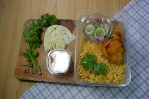 das braun arabisch Biryani kommt mit ein Hähnchen Bein im ein Plastik Box und verschiedene Seiten, einschließlich Ajhab, Gemüse, und Soße, alle auf ein Schneiden Tafel. foto