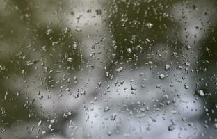 ein Foto von Regentropfen auf dem Fensterglas mit verschwommenem Blick auf die blühenden grünen Bäume. abstraktes Bild, das bewölkte und regnerische Wetterbedingungen zeigt