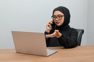Porträt von attraktiv asiatisch Hijab Frau Arbeiten auf ihr Laptop. Muslim Mädchen Herstellung ein Telefon Anruf im Büro. Mitarbeiter und freiberuflich Arbeiter Konzept. foto