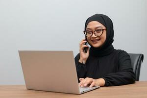 Porträt von attraktiv asiatisch Hijab Frau Arbeiten auf ihr Laptop. Muslim Mädchen Herstellung ein Telefon Anruf im Büro. Mitarbeiter und freiberuflich Arbeiter Konzept. foto