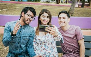 drei lächelnd freunde nehmen ein Selfie Sitzung im das Park, drei lächelnd freunde Sitzung im das Park nehmen ein Selfie. Vorderseite Aussicht von drei glücklich freunde nehmen ein Selfie während Sitzung auf ein Bank foto