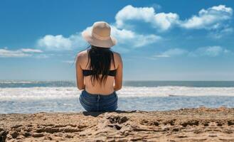 Mädchen im Hut Sitzung mit ihr zurück gegenüber das Meer, jung Frau im Hut Sitzung mit ihr zurück suchen beim das Meer, Ferien Mädchen Konzept, Rückseite Aussicht von ein Mädchen suchen beim das Meer foto