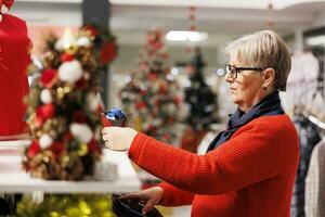 Senior Frau Surfen zum Zubehör und Mode Artikel im festlich Verkauf speichern, suchen zum Geschenke und Weihnachten Abendessen Outfit. Kunde Überprüfung Material von Kleidung auf Gestelle beim Einkaufen Einkaufszentrum. foto