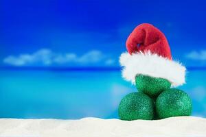 glänzend Grün Bälle im rot Santa klaus Hut auf Sand von Strand mit Meer. Weihnachten, Neu Jahr im heiß Länder. Kopieren Raum foto