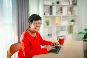 Senior alt asiatisch Frau Arbeiten nach Pensionierung mit Laptop beim heim. alt Freiberufler Arbeiten oder Lernen Neu Technologie auf Laptop im Leben Zimmer. Pensionierung Aktivität Konzept. foto