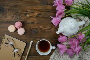auf ein hölzern Tabelle Dort ist ein Weiß Teekanne, ein Tasse von Tee, ein Notizbuch mit ein Rahmen von Gläser, und schön Tulpen. foto