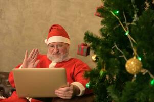 glücklich Santa claus mit Laptop winken Hand auf Video Berufung Kind reden zu Kind Gruß auf fröhlich Weihnachten und glücklich Neu Jahr beim Zuhause während Sitzung auf Couch Nächster zu dekoriert Weihnachten Baum foto