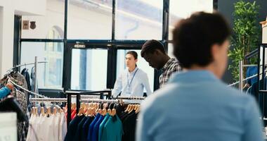 afrikanisch amerikanisch Kunde suchen beim Kleiderbügel mit modisch Shirt, Analysieren Kleider Stoff Vor Kauf Es. Shopaholic Mann Einkaufen zum modisch Fan-Shop und Zubehör im modern Boutique foto