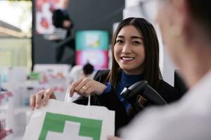 Drogerie lächelnd asiatisch Käufer halten Einkaufen Tasche mit Kauf und diskutieren Medikamente mit Apotheke Geschäft Arbeiter. Frau Kauf medizinisch Produkt beim Medikament Verkauf Geschäft Zähler Schreibtisch foto