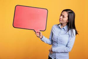 Filipino Frau halten leeren rot Rede Blase mit Kopieren Raum, Werbung Produkt. heiter jung Erwachsene Stehen im Studio Über Gelb Hintergrund, suchen beim leer Dialog Rahmen mit Platz zum Text foto