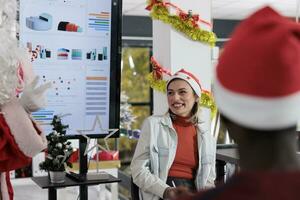 Geschäftsfrau haben Spaß während Geschäft Treffen im Weihnachten dekoriert Sitzungssaal während bezahlen Beachtung zu Öffentlichkeit Lautsprecher gekleidet wie Santa Klaus. Mitarbeiter Lachen, genießen korporativ Vorlesung foto