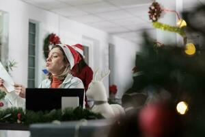 müde unzufrieden Mitarbeiter überwältigt durch stressig schwierig Papierkram im Weihnachten dekoriert Büro. Geschäftsfrau Leiden von Ausbrennen Arbeiten auf Anspruchsvoll Unternehmen Projekt während Weihnachten festlich Jahreszeit foto