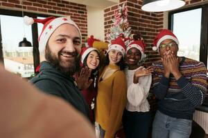 lächelnd Mann im Santa Hut nehmen slefie mit Kollegen im dekoriert Büro beim Weihnachten korporativ Party. Mitarbeiter suchen beim Kamera während posieren zum Handy, Mobiltelefon Telefon Gruppe Foto beim festlich Arbeitsplatz