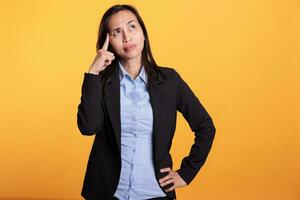 nachdenklich Filipino Frau halten Finger auf Tempel Brainstorming Ideen, posieren zweifelhaft im Studio Über Gelb Hintergrund. nachdenklich Geschäftsfrau im formal passen Nachdenken Über ernst Problem foto