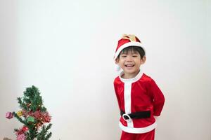 asiatisch Junge tragen Santa Kostüm Stehen und spielen in der Nähe von ein Spaß Weihnachten Baum auf Weiß Hintergrund. foto