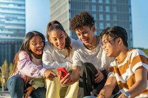 Gruppe von glücklich Teenager freunde suchen das Telefon und Lachen im ein Bank im das Stadt Straße. foto