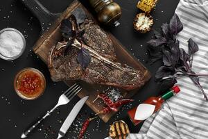 saftig gegrillt groß T-Bone Steak garniert mit Kräuter, Mais und Salz- mit Gabel und Messer neben es auf Schneiden Tafel foto