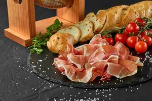 Jamon geschnitzt von Schwein Bein auf ein Jamonera Stand, Kirsche Tomaten, Stangenbrot, Kräuter, Gewürze auf schwarz Schiefer Tafel. dunkel Hintergrund. Nahansicht. Seite Sicht. foto