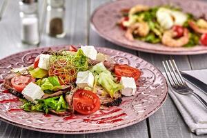 gesund gemischt griechisch Salat serviert auf ein Rosa Teller mit Silber Gabel enthält knackig belaubt grüne, mikrogrün, Feta, Zwiebel, Tomate und geschnitten Rindfleisch foto