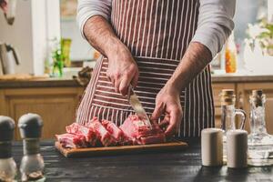 Mann schneidet von frisch Stück von Rindfleisch auf ein hölzern Schneiden Tafel im das Zuhause Küche foto