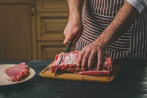 Mann schneidet von frisch Stück von Rindfleisch auf ein hölzern Schneiden Tafel im das Zuhause Küche foto