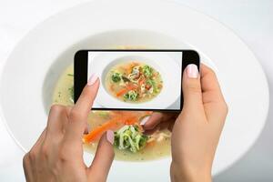 Schießen Essen auf Telefone Kamera. Hähnchen Suppe mit Brokkoli, Möhren und Sellerie im ein Weiß Schüssel auf ein Weiß Hintergrund. foto