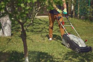 Fachmann Gärtner im beiläufig Outfit und Handschuhe ist Trimmen Grün Gras mit modern Rasen Mäher auf seine Hinterhof. Gartenarbeit Pflege Ausrüstung. sonnig Tag foto