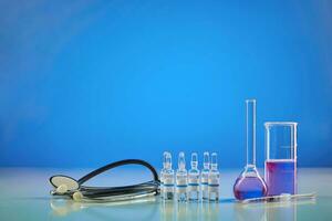 Messung Becherglas, medizinisch Flasche mit lila chemisch Reagens. sechs Ampullen mit flüssig, Spritze und Phonendoskop, Blau Hintergrund. Pandemie covid-19 foto