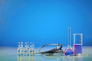 Becherglas, medizinisch Flasche mit lila chemisch Reagens. sechs Ampullen mit flüssig, Spritze und Phonendoskop, Blau Hintergrund. weltweit Pandemie covid-19 foto
