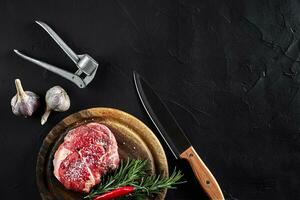 Stück von Rindfleisch Filet, mit Messer zum Schneiden und Hacken Fleisch, Gewürze wurden Kochen - - Rosmarin, Pfeffer, Salz, Knoblauch. auf alt hölzern Tafel auf Stein schwarz Tisch. foto