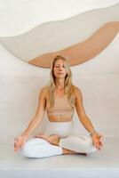 Innen- Foto von jung blond Frau tun Yoga Übungen und üben Meditation. Sitzung im Lotos Pose im Yoga Klasse.