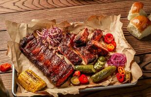 gegrillt Schweinefleisch Rippen serviert mit gegrillt Mais, Salat, Grill Soße, Salz- Pfeffer und Gurke auf Pergament Papier auf ein hölzern Tisch. oben Aussicht foto