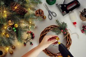 weiblich Hände schmücken das Weihnachten Kranz mit Fichte Geäst mit ein rot Beere und Wald Zapfen. Weiß Hintergrund. Neu Jahre Konzept. foto