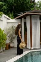 schön Modell- Frau im modisch Sonnenbrillen und schwarz Sommer- Outfit mit Stroh Hut posieren auf modern Villa. voll Länge. foto