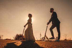 Silhouette Hochzeitspaar, das bei Sonnenuntergang auf Rollern entlang der Straße fährt foto