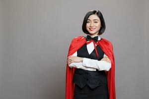 lächelnd zuversichtlich asiatisch Kellnerin im rot Superfrau Mantel Stehen mit Waffen gekreuzt Porträt. heiter Frau Rezeptionist mit gefaltet Hände tragen Held Kap suchen beim Kamera foto