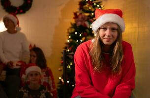 Porträt von Teenager Mädchen im Santa Hut Sitzung auf Stuhl im Vorderseite von Weihnachten Baum, fröhlich Weihnachten und glücklich Ferien foto